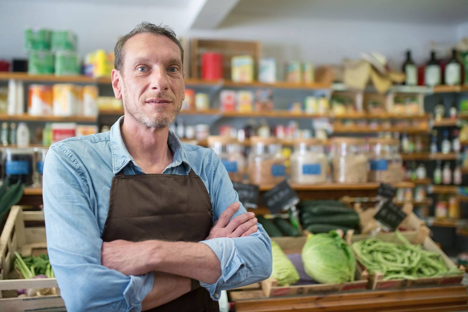 Un épicier en tablier, bras croisés, devant ses étals de légumes et ses rayons.