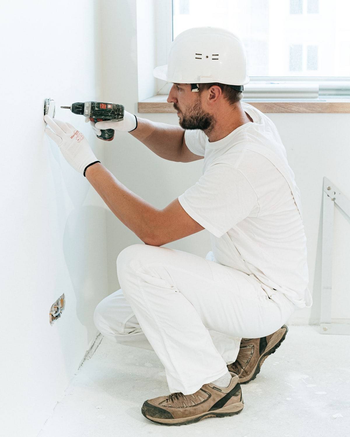 Un électricien casqué, accroupi, perce le mur au-dessus d'un boîtier électrique.