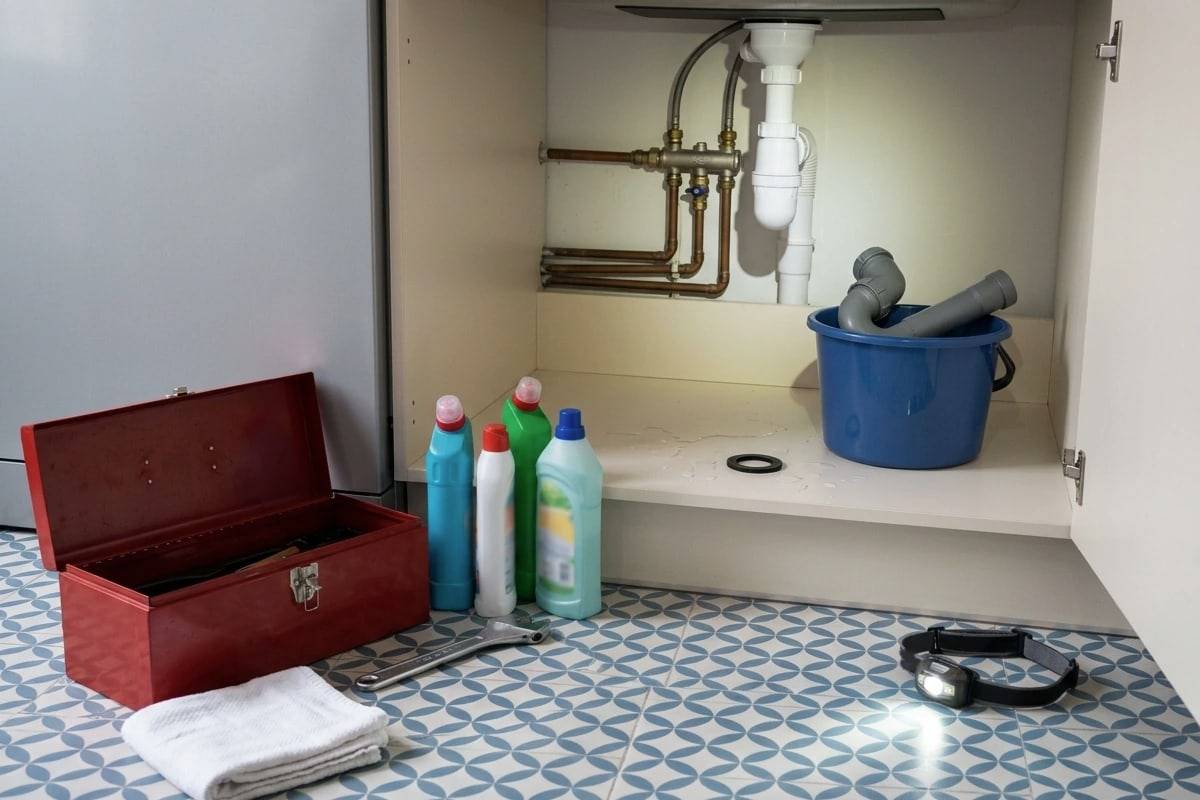 Sous un évier ouvert, une caisse à outils rouge, une lampe frontale et un seau, sur un sol en carreaux de ciment.