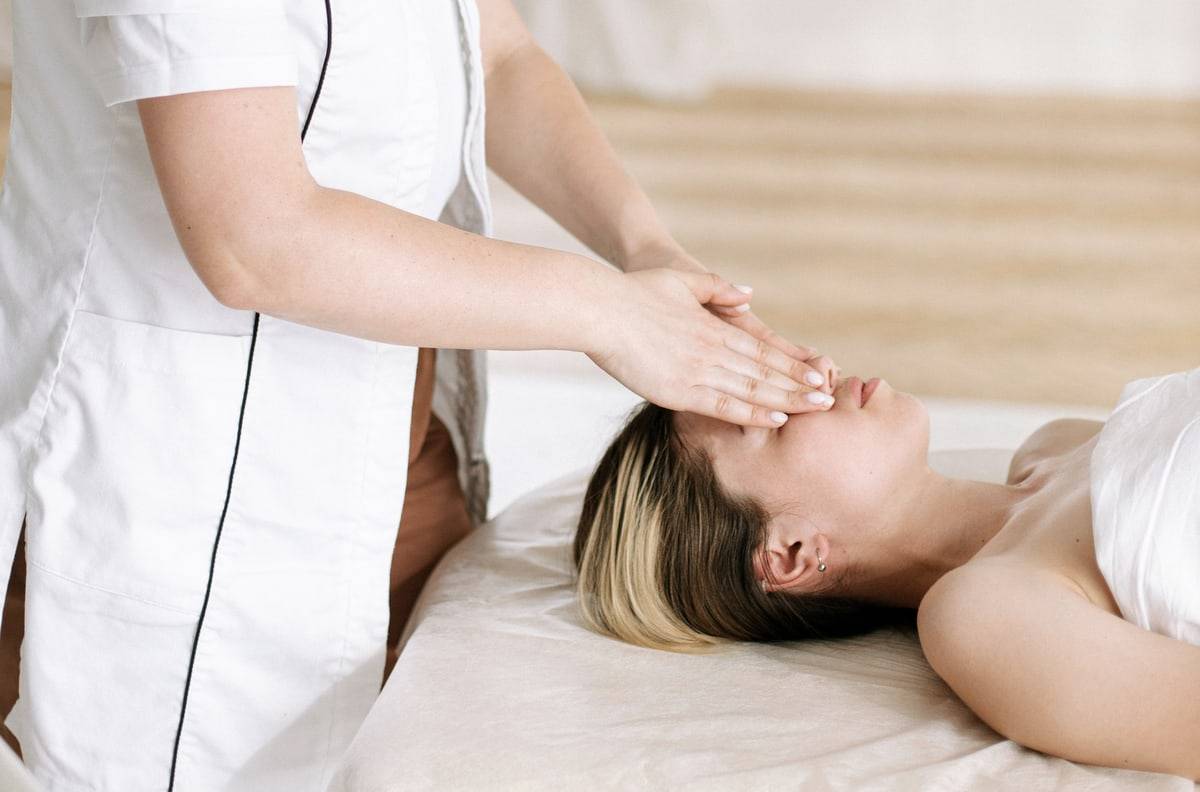 Une praticienne en blouse blanche pose les mains sur le front d'une cliente allongée.