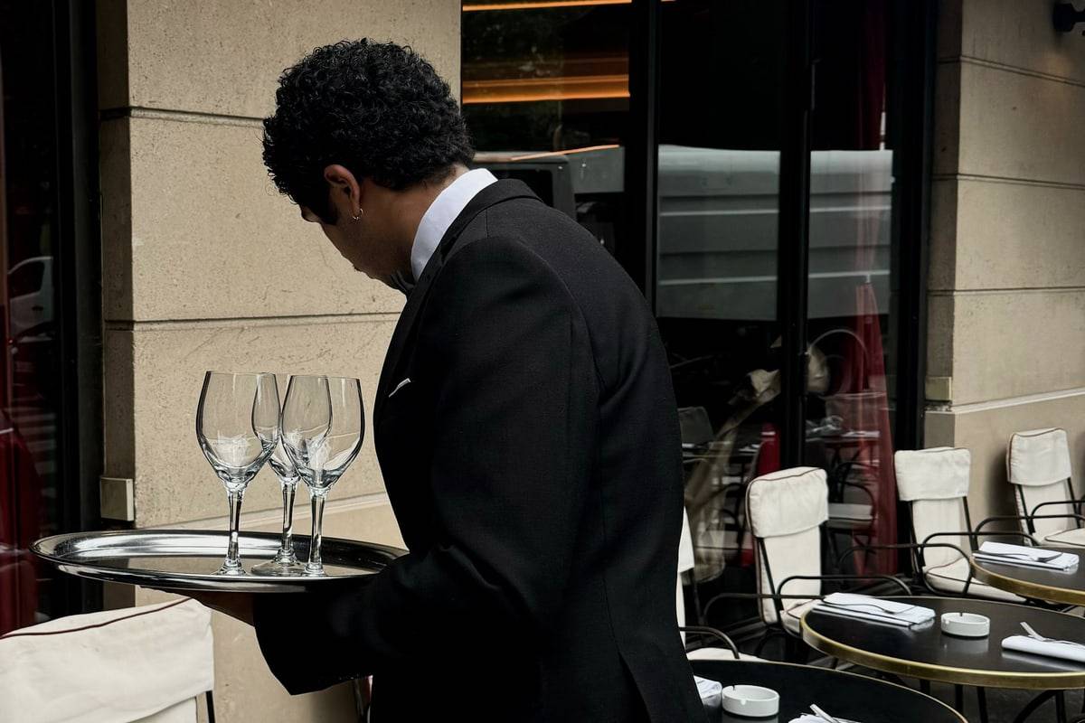 Un serveur en veste noire porte un plateau de verres à pied entre les tables rondes d'une terrasse.