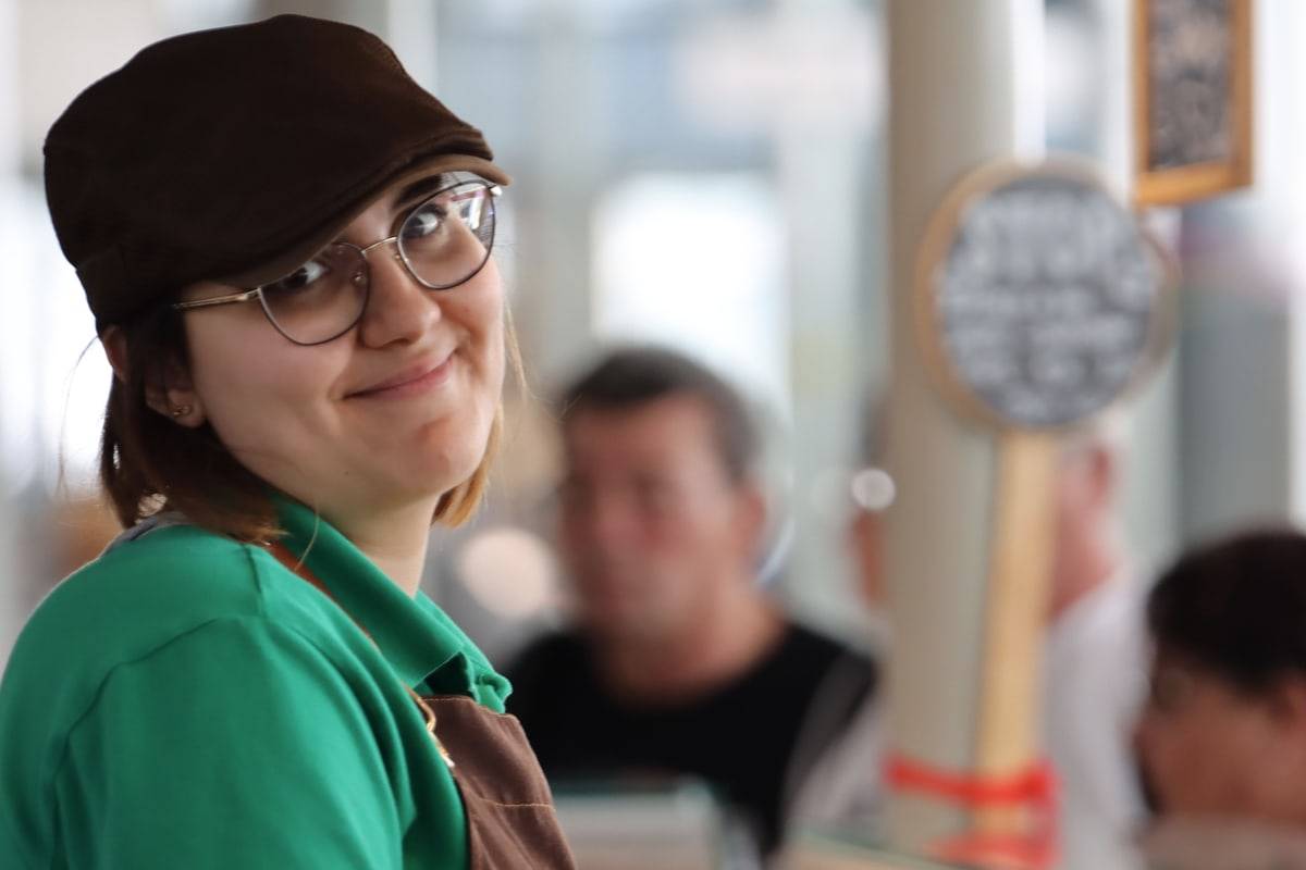 Une fromagère en casquette et lunettes sourit, tournée vers l'objectif, derrière son comptoir.